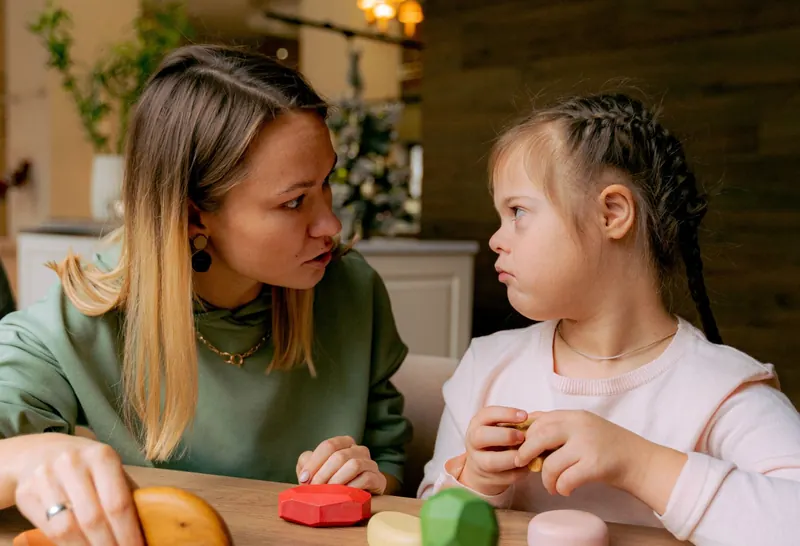 An adult and a girl sit facing each other at a wooden table with colorful wooden toys