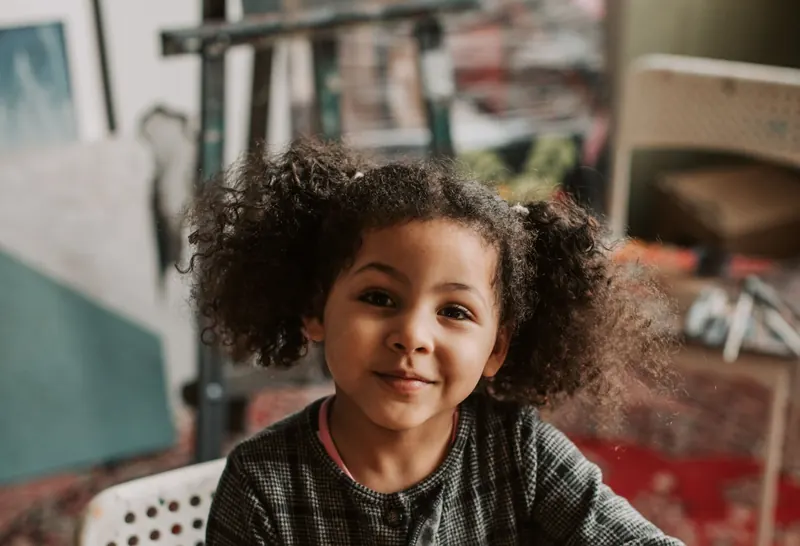 A smiling girl with curly hair sits at a craft table playing with modeling clay
