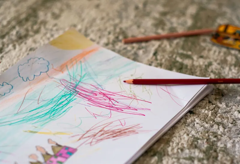 A child's colorful scribble drawing on paper with a red pencil resting on top