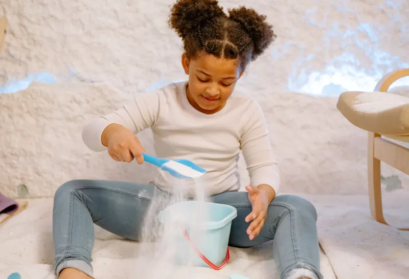 A smiling girl sits barefoot on a soft white floor, scooping fine white granules into a small blue bucket